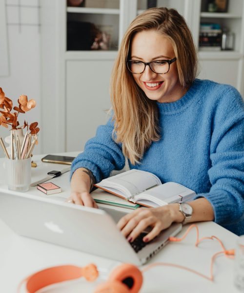 A cheerful woman in a blue sweater working remotely with a laptop in a cozy home setting.